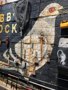 This piece is 'Cobb Dock & The Red Hook Lady Fliers', 2017. It is made of wood, tin, roofing tar, latex paint, slipcast ceramic pigeons and performance ephemera from "Fly By Night". It includes a large mural painting and shows the skinned facade and side of a coop from the "Fly By Night" performance.