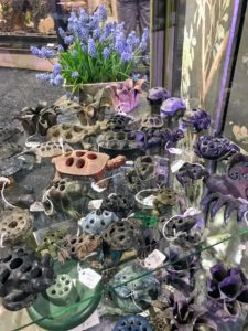 Here is a table filled with all kinds of flower frogs. Flower frogs are made of lead, pottery, glass, or bronze and sit at the bottom of a bowl or a vase to hold flower arrangements firmly in place.
