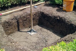 The team adjusts the hole and using a steel tamper, packs the soil firmly, so it is level. The hole is about 23-inches deep.
