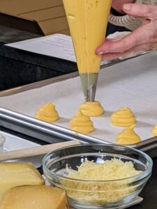 These are made with water, butter, sugar, salt, flour, eggs, Gruyere cheese, and Compte cheese and then transferred to a pastry bag fitted with a half-inch plain tip. Here I am piping the batter onto a parchment paper-lined baking sheet. Each mound is about an inch in diameter.
