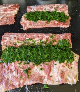 The lamb saddle is a cut comprising the racks that are deboned but still held together by the skin. First, Chef Pierre cuts a lot of the fat off and the meat is flattened with a meat tenderizer. The deboned rack of lamb is placed, skin-side down, on a board and then the spinach and herb mixture is spread over the meat.