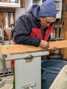 One of the most important rules in carpentry is to double-check one's measurements for accuracy before cutting a piece of wood. Here, Pete makes sure he cuts the wood to match the existing pieces of the stand.