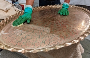 The next step is to clean the center of the tray. Carlos rubs more cream into the piece using the sponge. Whenever polishing metals, be sure you use a product that is specifically made for the type of metal being cleaned.