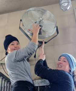 Mountable models are positioned above each stall where they can be safely secured and plugged into outlets. Helen and Dolma install this new unit in a stall that did not have a fan.