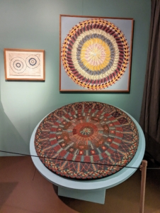The "Knit Wool Rug" in the foreground was made by Elvira Curtis Hulett when she was 88-years old living with Shakers in Massachusetts. It shows her attention to detail as well as the utilitarian functionality so often shown in Shaker pieces. Above it on the wall is a more contemporary work by Miriam Schapiro called "Golden Pinwheel" which was made using braided and knitted wool.