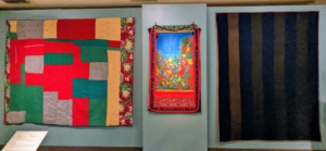On the left is a quilt by renowned African American quilter, Mozell Stephens Benson in 1991. The center piece is Faith Ringgold's "Feminist Series: Of My Two Handicaps #10." Faith is known for popularizing the story quilt, a quilt with pictures and textures used primarily to tell a story. On the right is "Bars Quilt" made by Amish artist Elizabeth Yoder between 1870 and 1880.