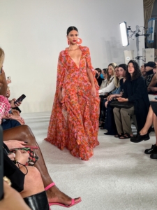 This dress shows off a flowing cape and deep v-neckline. The model accessorizes with large salmon colored matching earrings.