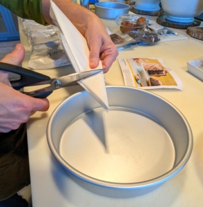 While the oven is pre-heated to 300-degrees Fahrenheit, Sarah Carey prepares the eight-inch baking pans with parchment rounds. She folds a piece of parchment paper in half, then in half again, folds it into a triangle twice and holds the folded paper over the pan with the center point over the center of the pan. Then she cuts the parchment using the outer edge of the pan as her guide.