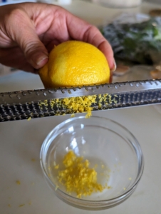 The first step is to zest all the lemon. Elvira uses a microplane and runs the lemon up and down over the surface, scraping off the yellow skin into the bowl. Lemon zest, or the zest of any citrus fruit, refers to the outermost layer of the peel known as the flavedo. This layer contains natural oils that are full of sweet citrus flavor.