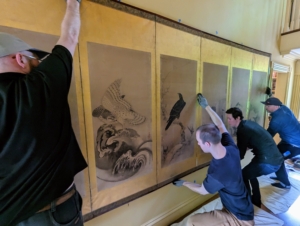 The first one to go up is this screen in the foyer. All four gently pick it up and put it in place. The team always wears gloves while handling precious art work, so there are no finger prints left behind.