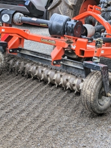 Here is a closer look at the roller of the power rake. When it is lowered onto the road surface and tilted to the proper angle, this attachment moves the gravel and road dust to the center, creating the proper crown for the surface. There should be about a three-percent slope from the shoulder to the center of the road. On less used roads, the power rake freshens up the existing gravel as it turns and brings any compacted gravel to the surface.