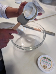 The first step is to empty the two tubs of mascarpone into a medium bowl and combine with 1/3-cup water. Mascarpone is made similarly to American cream cheese, but it uses a base of whole cream rather than milk. Like cream cheese, it is a fresh cheese that is not aged before it is eaten.