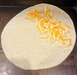 A spoonful of cheese is placed on one side of the tortilla. Brian prepares these on a rimmed baking sheet, so they are ready to put into the oven.