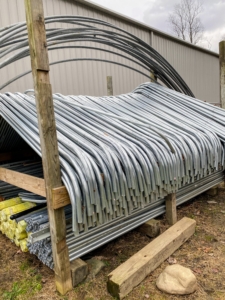 These pipes, which we keep organized in a back field where we store other supplies, are made from rolled galvanized 16 gauge industrial tubing. They are part of the framing supplies needed for our protective burlap covers. In all, hundreds of ground stakes, purlin pipes, connectors, nuts, and bolts are used for the project. They are actually the metal parts of greenhouse hoop houses.
