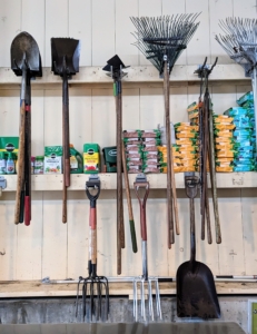 Rakes, forks, edgers, hoes, and other tools are also hung in the same area.