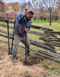 Pete already removed the visible section of post, but it broke in two and the bottom is still in the hole. He tries to loosen the soil around it by hand using the tamping bar.