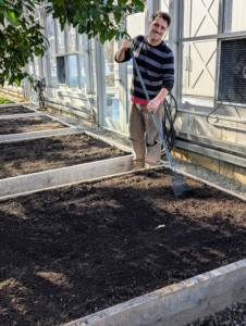 This greenhouse has 16 of these wooden garden boxes to fit the entire length and width of the space. One larger bed is at one end. Raised bed gardening allows good drainage, prevents soil compaction, and provides protection for those plants that may otherwise get trampled. Ryan starts by smoothing out the soil and wedding where necessary.