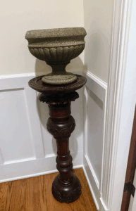 This is a decorative pedestal stand crafted from richly stained wood, showcasing detailed carved accents along its base and column. It supports an antique-style urn planter with a weathered, crackle finish.