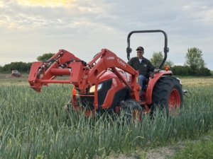 Keene practices foliar feeding, which is a method of applying nutrients directly to the leaves of the garlic plants. Here is Keene spraying each row of garlic. Foliar feeding allows for fast absorption, boosts plant growth, and increases the bulb size.