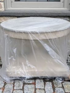 Because stone, cement, and clay-based materials can crack from exposure to the winter elements, it’s important to protect them from all sides. The first thing I do is cover the large opening with a piece of plywood. The crew cuts any spare plywood into rounds to fit over various pieces. Then it is wrapped in a layer of plastic all the way around.