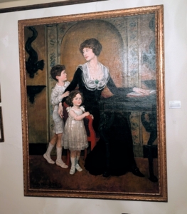 This is a family portrait of Lucie Bigelow Rosen and her children, Walter and Anne. This was painted in their New York City townhouse and is now displayed at Caramoor along with the green desk in the painting.